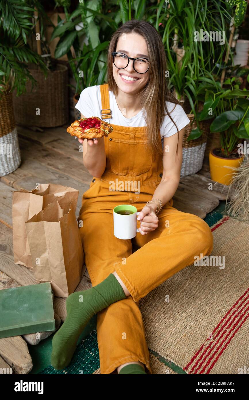 Donna sorridente fiorista/giardiniere in occhiali che indossano tute, seduta su un pavimento in legno nel suo giardino di casa, bere il tè, ha un buon umore per mangiare delicio Foto Stock