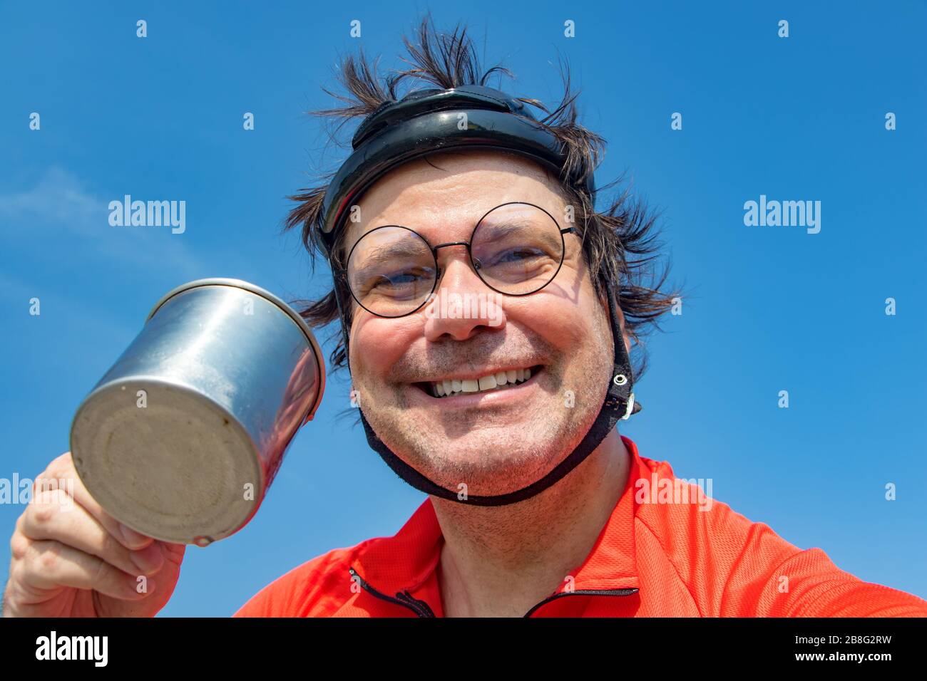 Ritratto di un ciclista sorridente in casco retrò beve acqua dal teacup. Bevanda dissetata del biker contro il cielo blu. Foto Stock