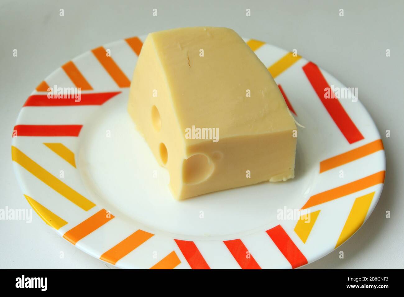 Un pezzo di formaggio giallo con buchi giace su un piatto bianco con un bordo colorato. Foto Stock