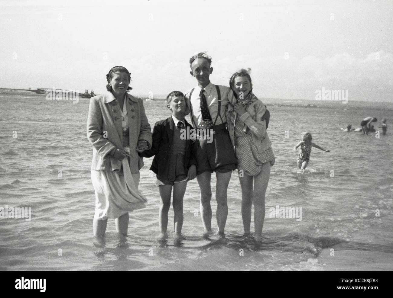 Anni '50, storico, una famiglia britannica con i loro pantaloni e abiti  arrotolati, posa per una foto in piedi in acqua di mare poco profonda al  bordo della spiaggia, Inghilterra, Regno Unito