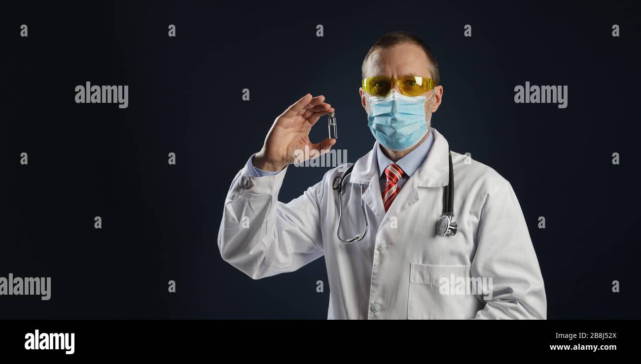 Medico maturo di sesso maschile che tiene un vaccino nelle sue mani. Il medico in una maschera medica protettiva e occhiali di protezione. Foto Stock