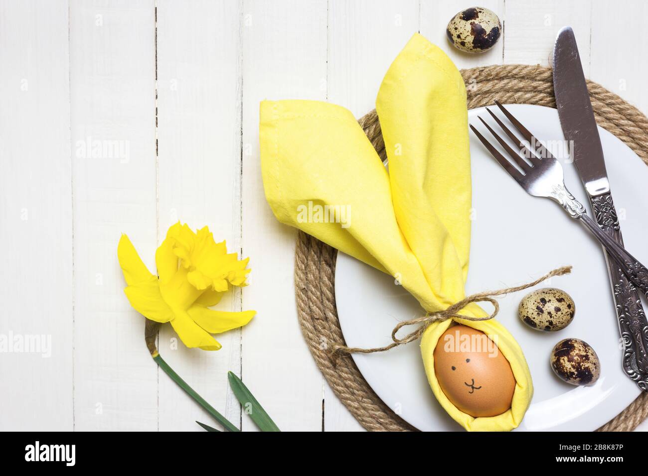 Bella festa tavola di Pasqua impostazione con uovo in tovagliolo come coniglietto di Pasqua e fiori di primavera su tavolo di legno bianco. Vista dall'alto con spazio di copia. Foto Stock