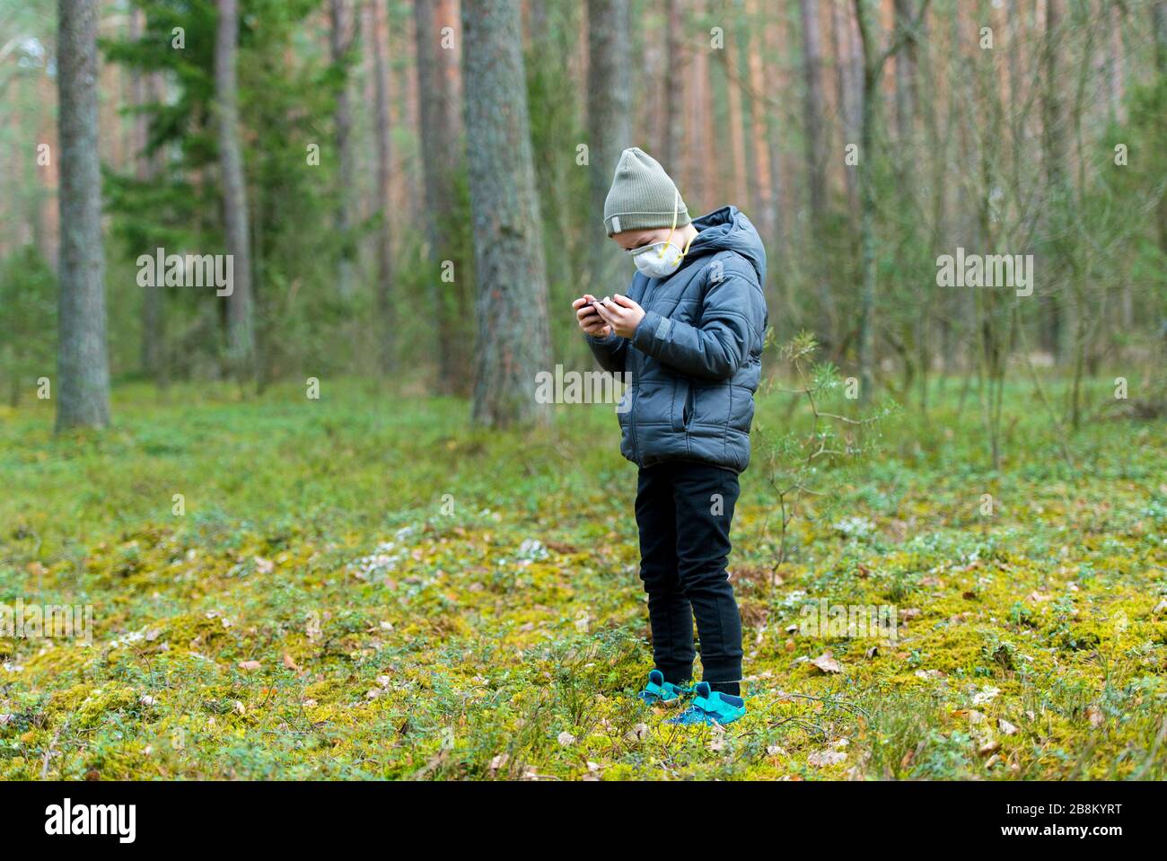 Capretto con maschera in bocca a causa della malattia di coronavirus e giocare i giochi mobili su un telefono nero in una foresta Foto Stock