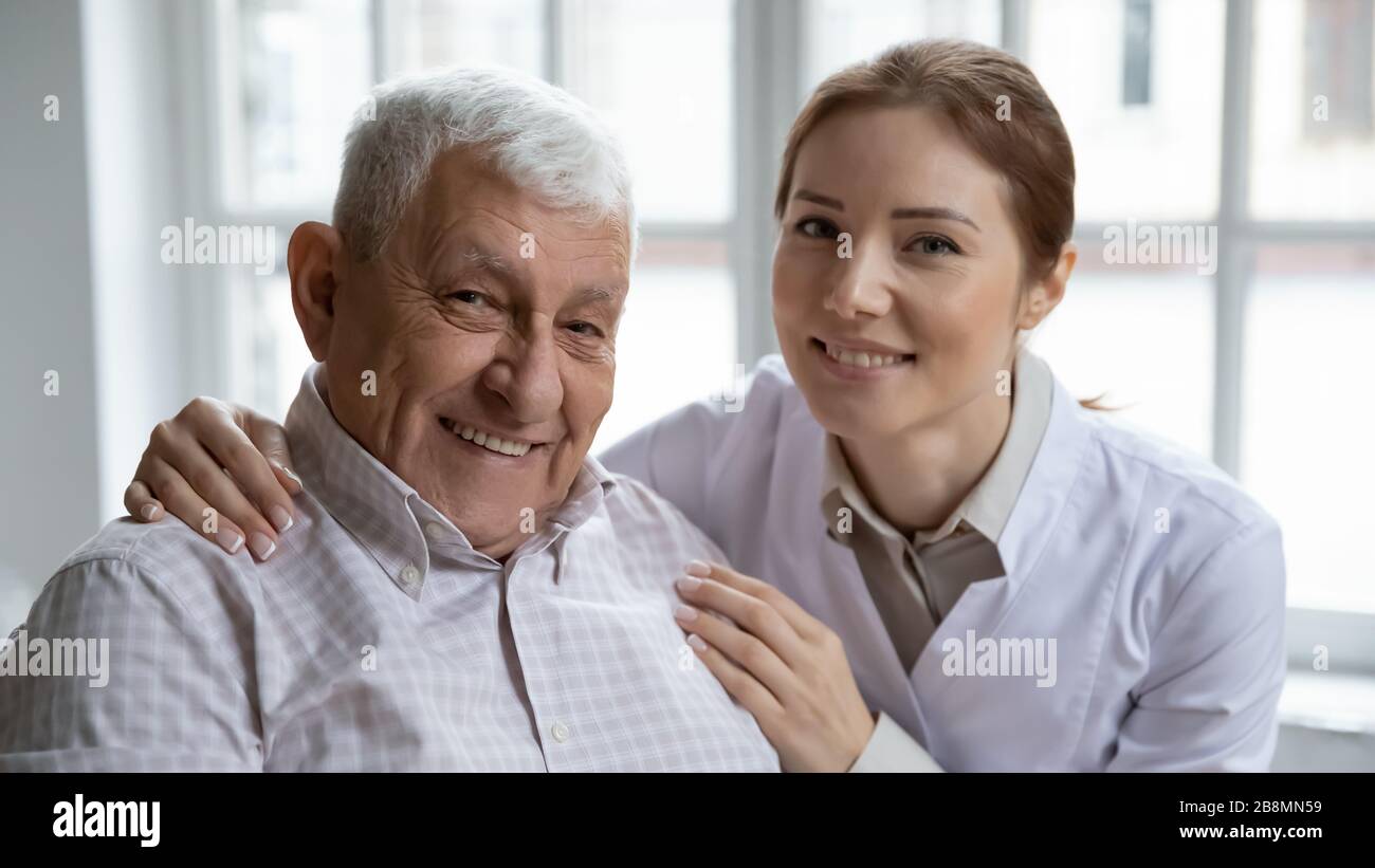 L'infermiera che si prende cura abbraccia il vecchio paziente sorridendo guardando la macchina fotografica Foto Stock