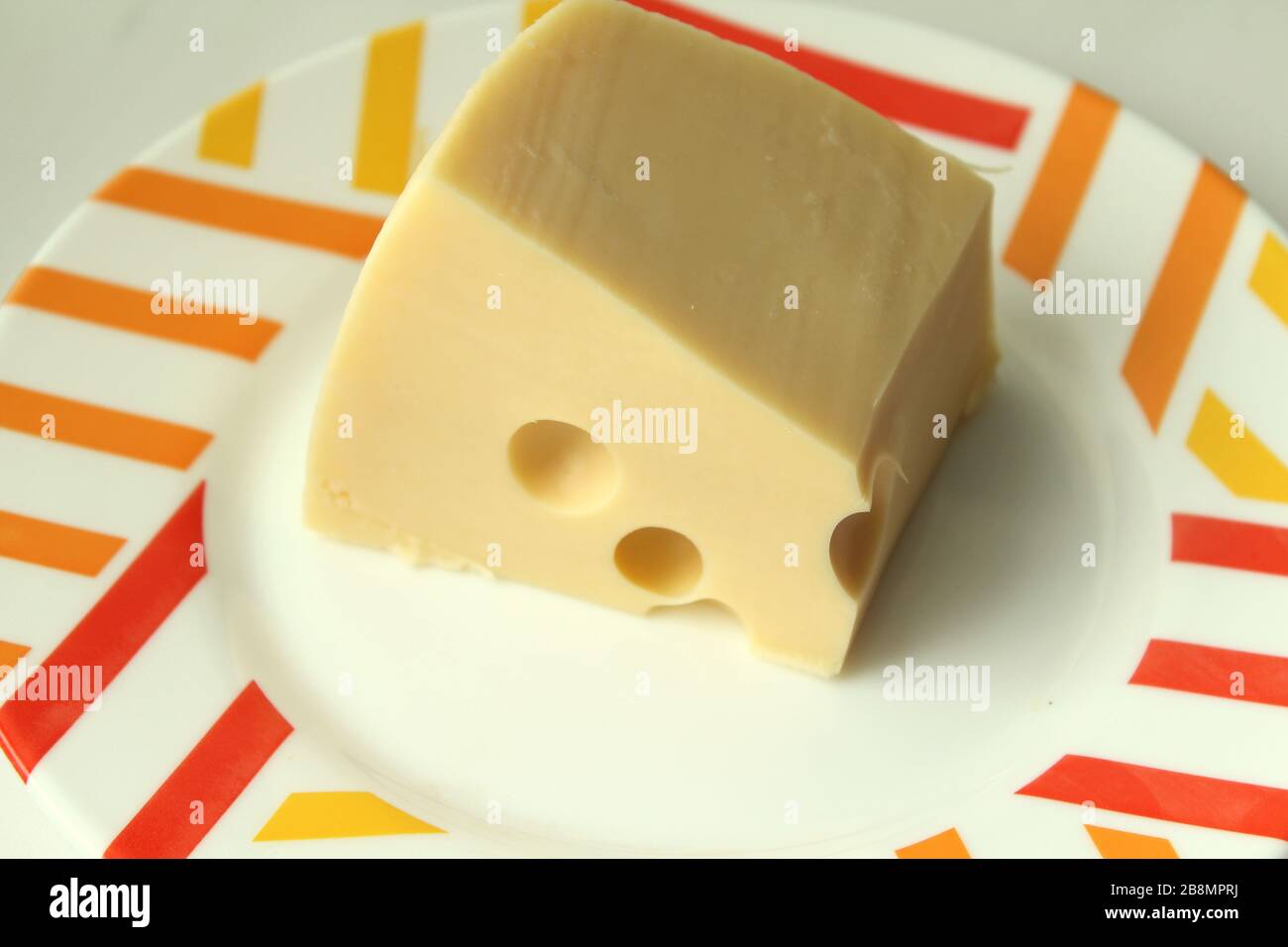 Un pezzo di formaggio giallo con buchi giace su un piatto bianco con un bordo colorato. Foto Stock
