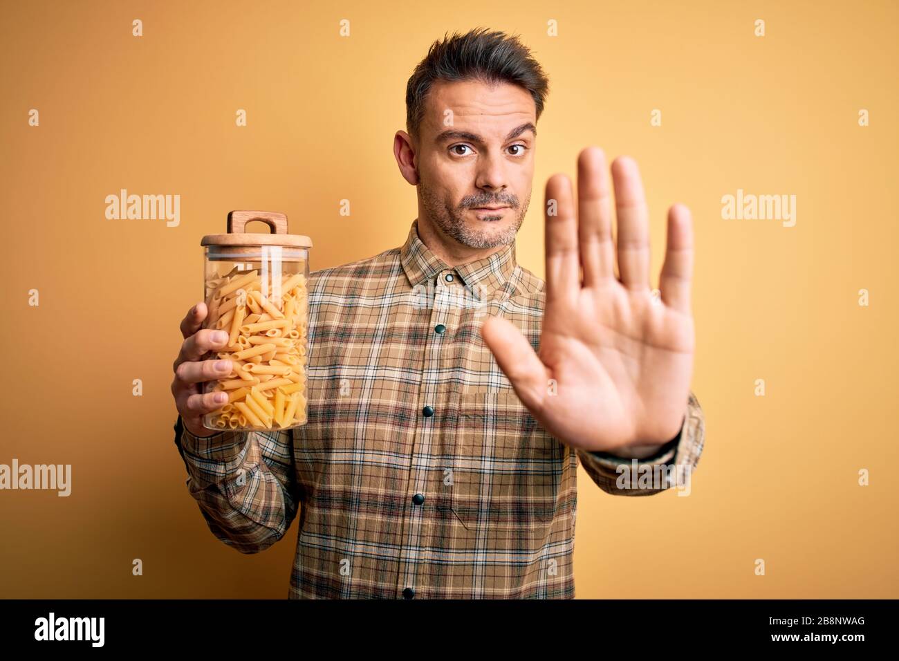 Giovane bel vasetto da uomo con pasta secca italiana maccheroni su sfondo giallo con mano aperta facendo stop segno con serie e fiduciosi expres Foto Stock