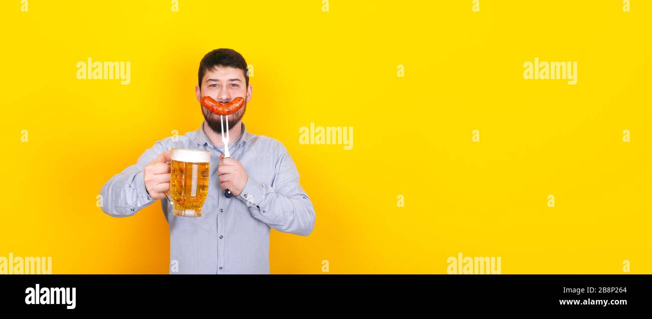 uomo con un bicchiere di birra e salsiccia alla griglia su una forchetta in mano, in piedi su sfondo giallo, mock-up panoramico Foto Stock