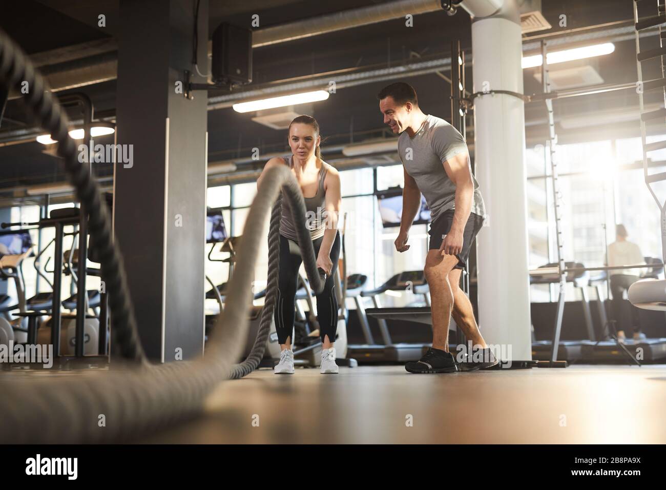 Ritratto grandangolare a lunghezza intera della giovane donna che fa esercizi di corda da battaglia durante l'allenamento trasversale in palestra moderna con allenatore personale, spazio copia Foto Stock