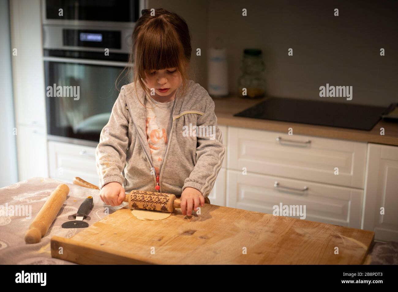 Bambina di 3 anni, in cucina bianca, spianatura pasta pizza con un piccolo perno di rotolamento su una tavola di legno. Idea dell'attività di blocco. Foto Stock