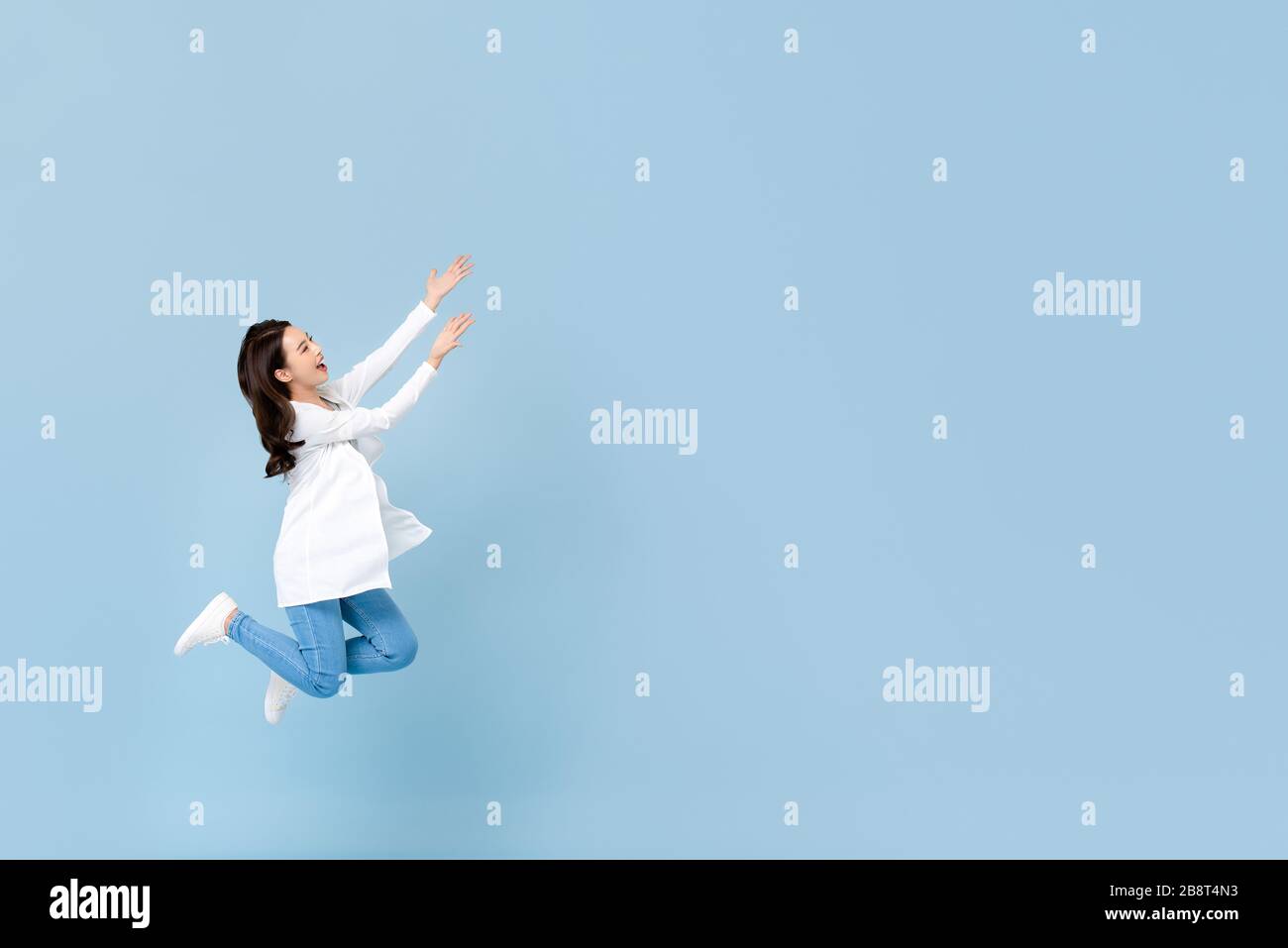 Ritratto a tutta lunghezza di giovane donna felice asiatica che galleggia a metà aria con le mani che si aprono allo spazio vuoto accanto in blu isolato studio sfondo Foto Stock