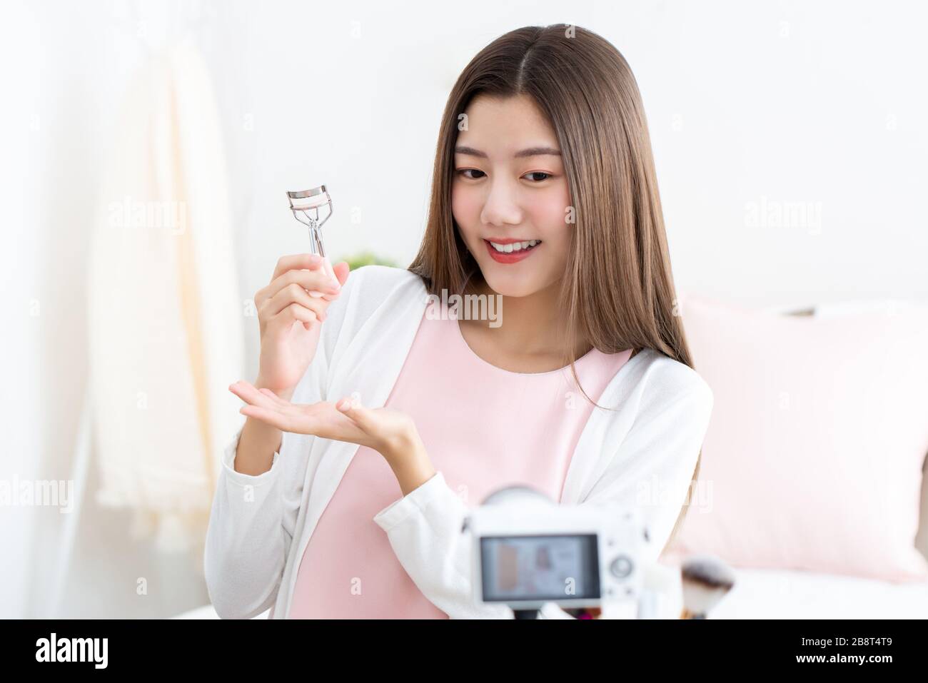 Giovane attraente donna asiatica influencer di bellezza che tiene arricciatore ciglia in streaming dal vivo davanti alla fotocamera Foto Stock