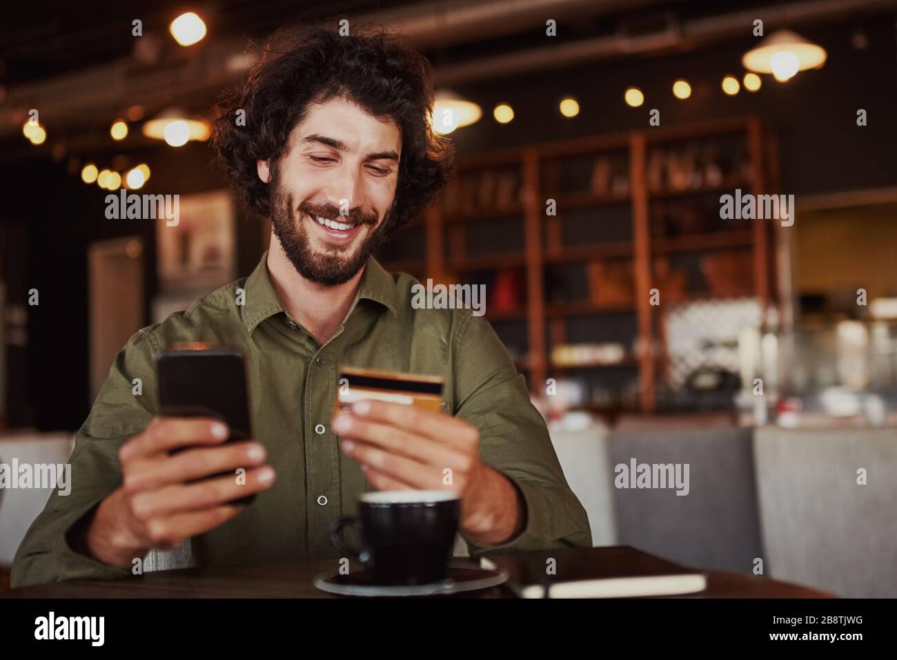 Felice caucasico bearded uomo che tiene il telefono cellulare digitando i dati della carta per effettuare il pagamento online seduto in caffetteria Foto Stock