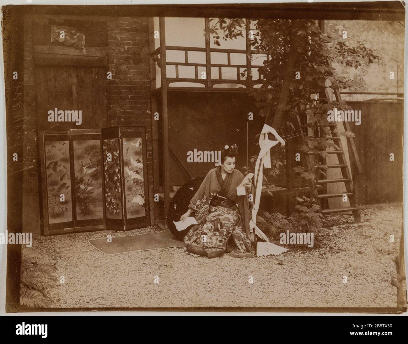 Giovane attrice in kimono che sbatte leggendo fuori davanti a uno schermo. Jeune actrice en kimono accroupie en train de lire à l'extérieur devant un paravent. Anonima fotografa, vers 1900. Parigi, musée Carnavalet. Foto Stock