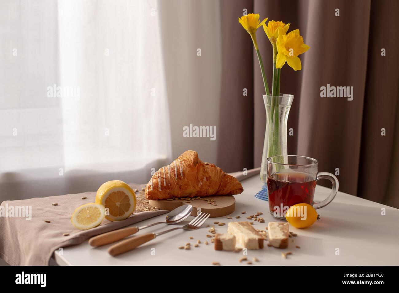 Nella sala sul tavolo accanto alla finestra con tende bianche e marroni, la colazione viene servita con una tazza di tè, croissant, limoni e narcisi gialle i. Foto Stock