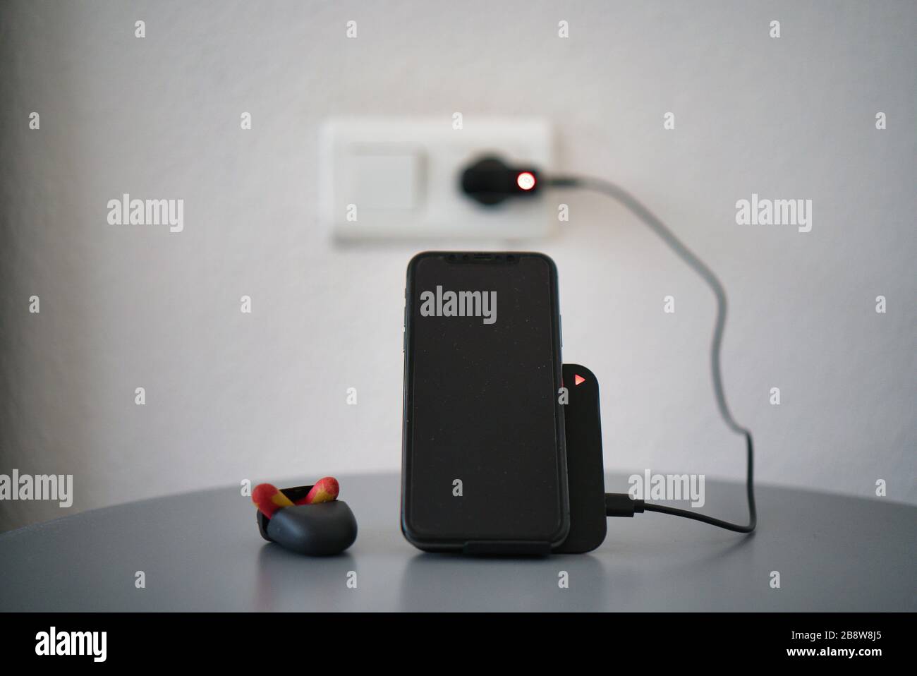 Caricabatterie wireless nero da tavolo con LED di ricarica per ricaricare uno smartphone. Display nero. Tavolo grigio argento. Tappi per le orecchie sul comodino Foto Stock