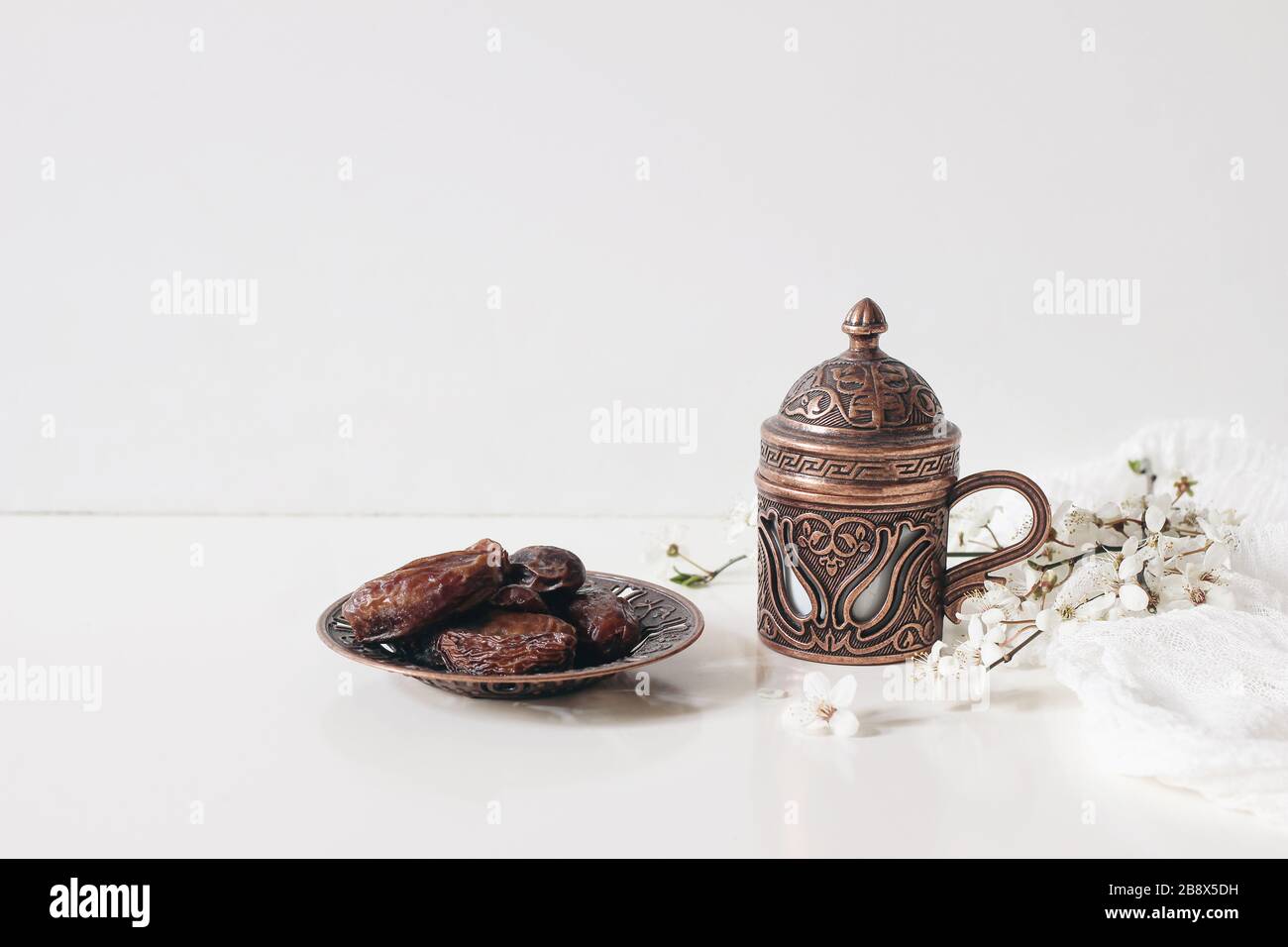 Biglietto d'auguri Ramadan Kareem, invito. Tè ornamentale, tazza di caffè, piatto di bronzo con frutta datteri, ramo di prunus bianco fiore su tavolo bianco. Foto Stock