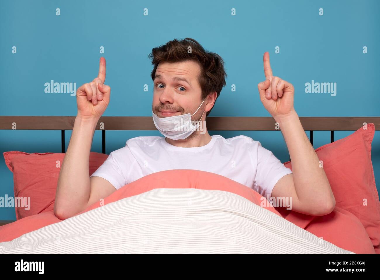 Uomo caucasico in maschera medica che riposa a letto su quarantena puntando qualcosa contro sfondo blu Foto Stock