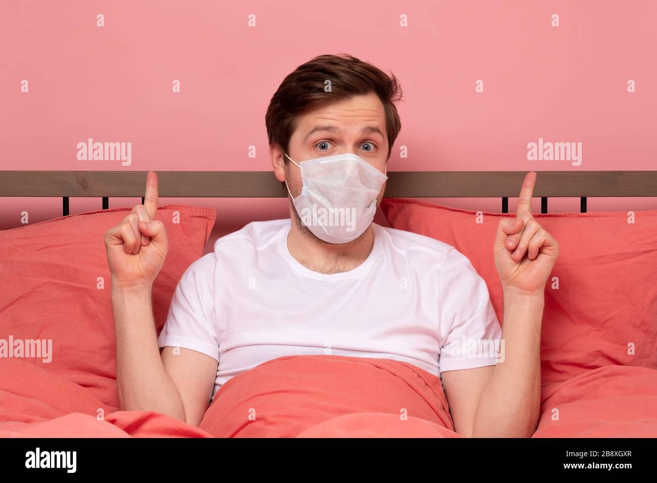 Uomo caucasico in maschera medica che riposa a letto su quarantena puntando qualcosa contro lo sfondo rosa Foto Stock