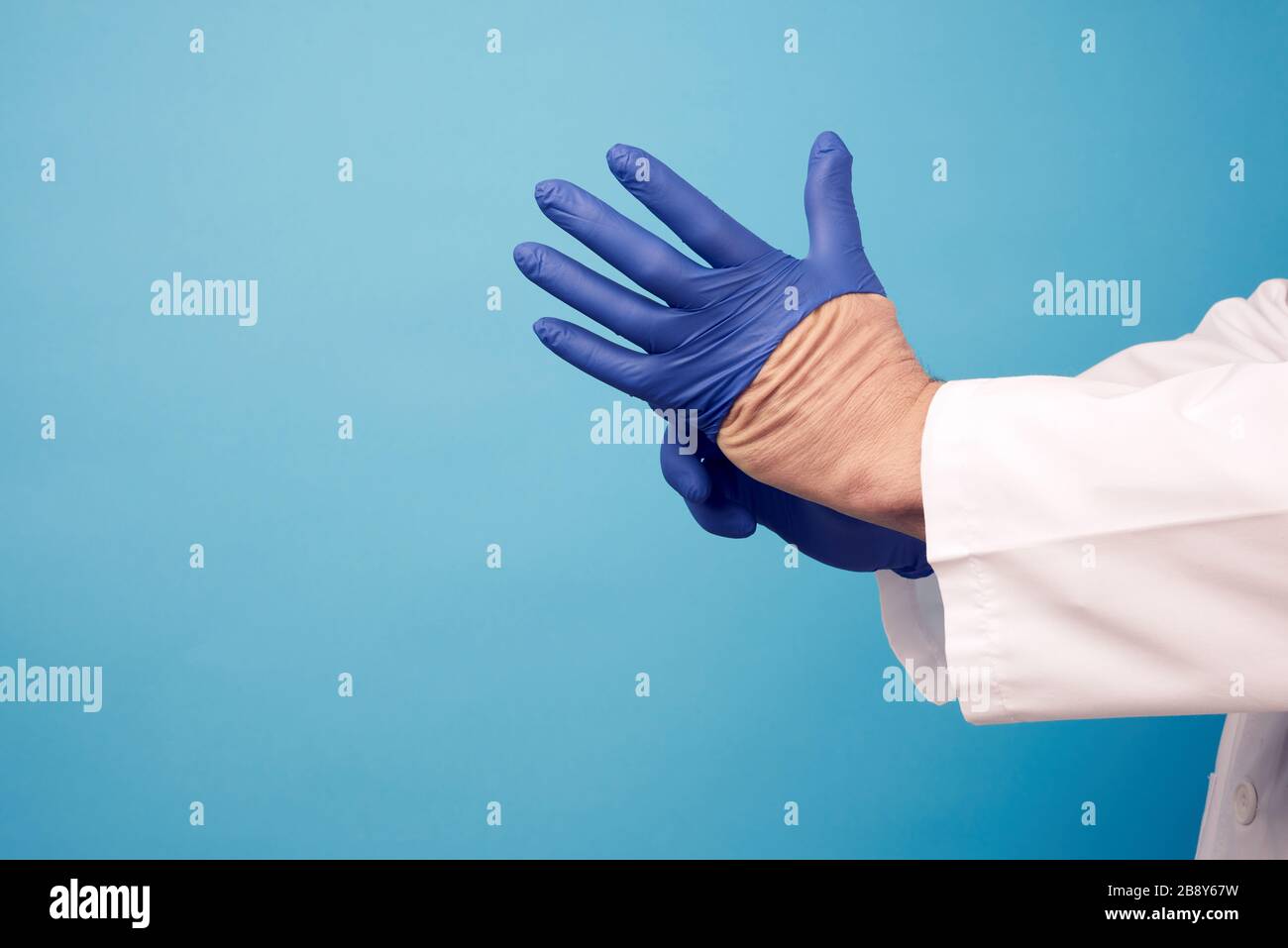 medico maschile in uniforme bianca mette sulle sue mani guanti in lattice blu sterile prima dell'intervento chirurgico, sfondo blu, spazio copia Foto Stock