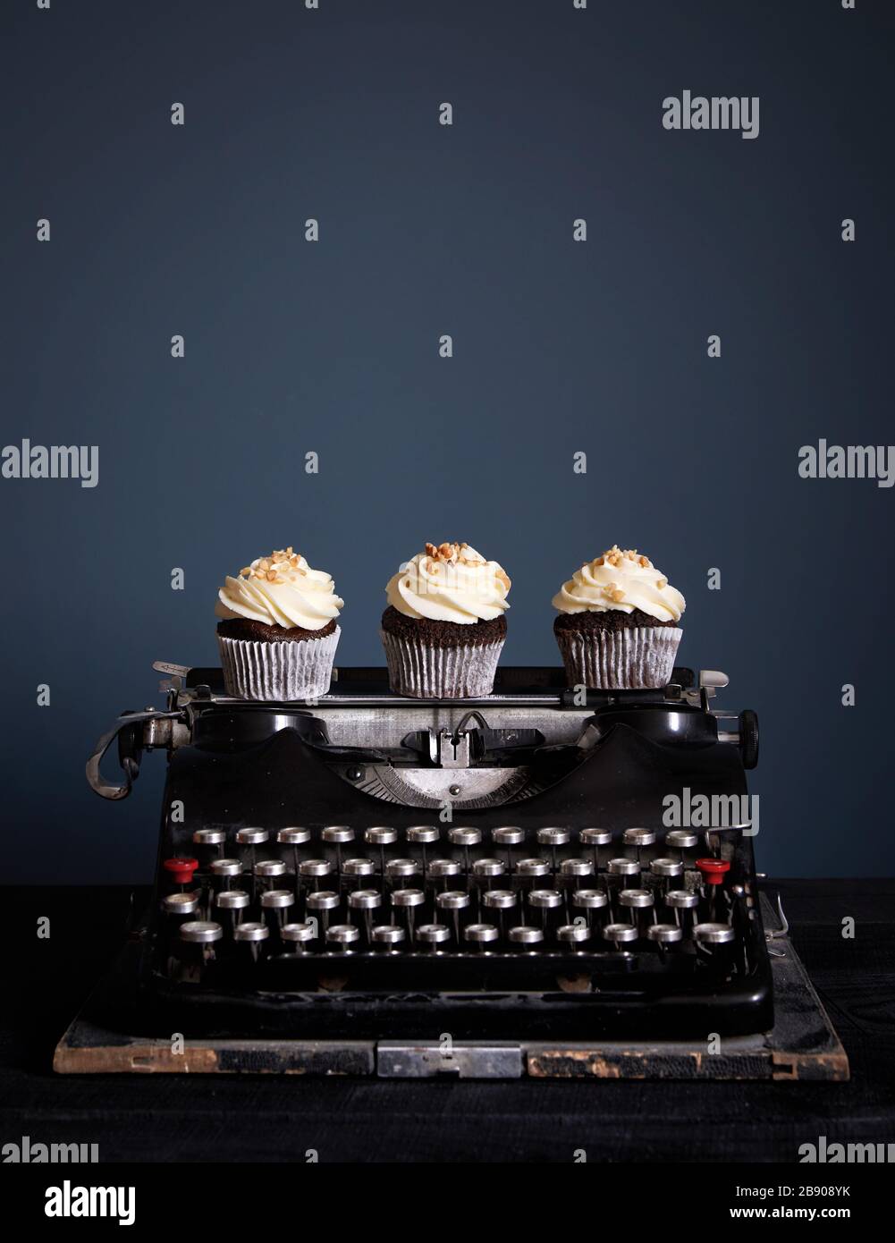 Cupcake al cioccolato decorati con crema bianca in piedi su vecchia macchina da scrivere vintage su sfondo scuro. Spazio libero per il testo Foto Stock