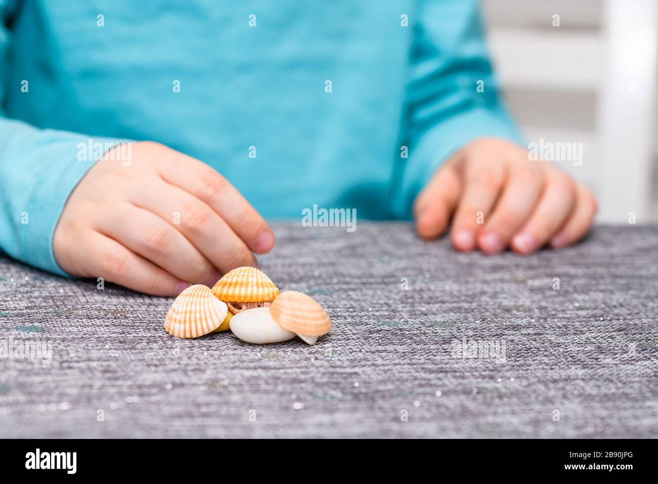 Primo piano di un ragazzo di cinque anni con un mucchio di conchiglie e ciottoli su un tavolo davanti a lui. Ha una camicia turchese. Foto Stock
