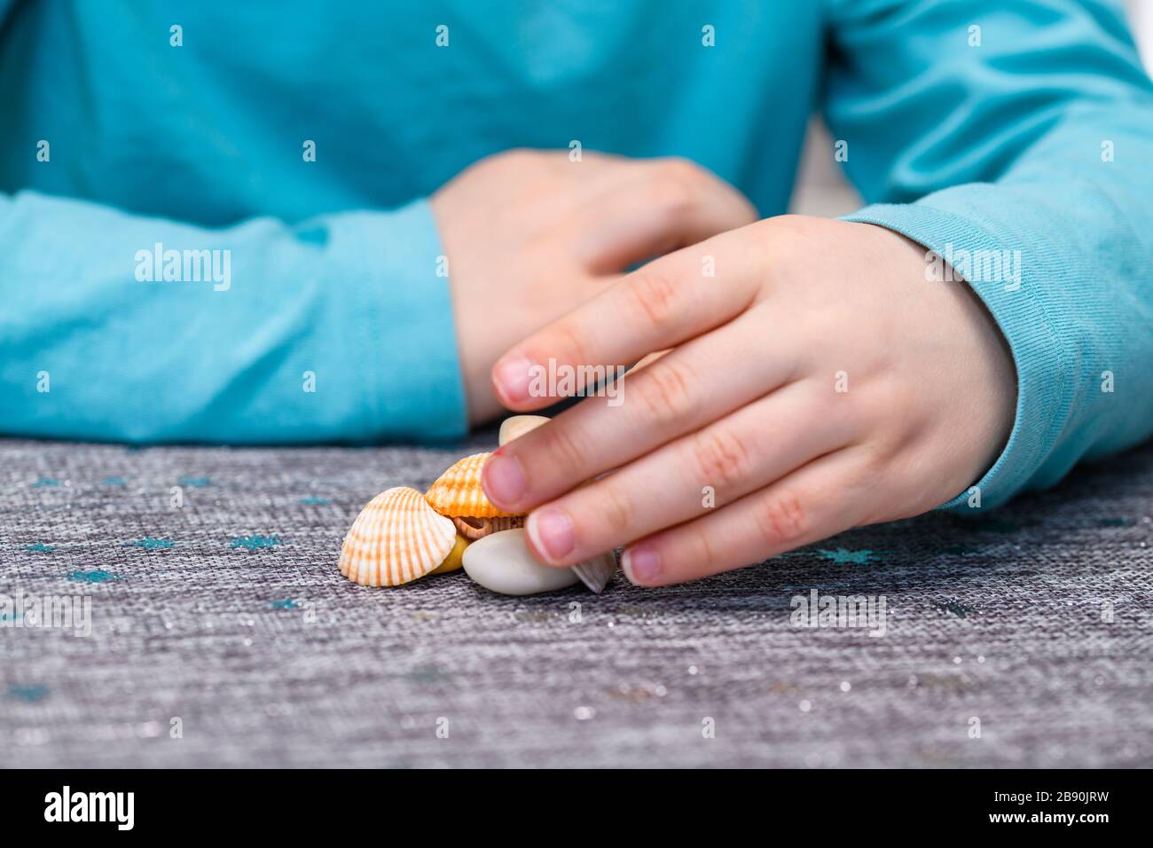 Primo piano della mano di un ragazzo di cinque anni che gioca con un mucchio di conchiglie e ciottoli su un tavolo. Ha una camicia turchese. Foto Stock