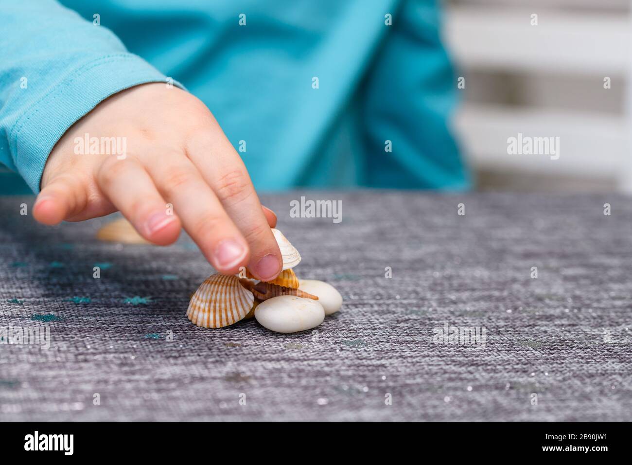 Primo piano della mano di un ragazzo di cinque anni che gioca con conchiglie e ciottoli su un tavolo. Ha una camicia turchese e raccogliendo un conchiglia. Foto Stock