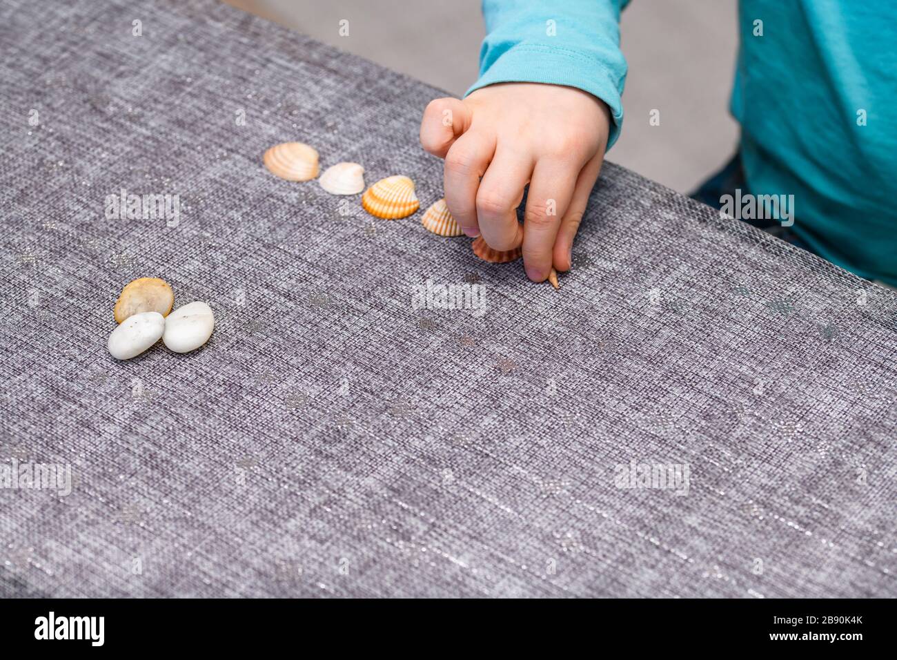 Ragazzo di cinque anni che si allineano su conchiglie e ciottoli su un tavolo. Girato dall'alto, ha una camicia turchese e raccoglie un conchiglia. Foto Stock
