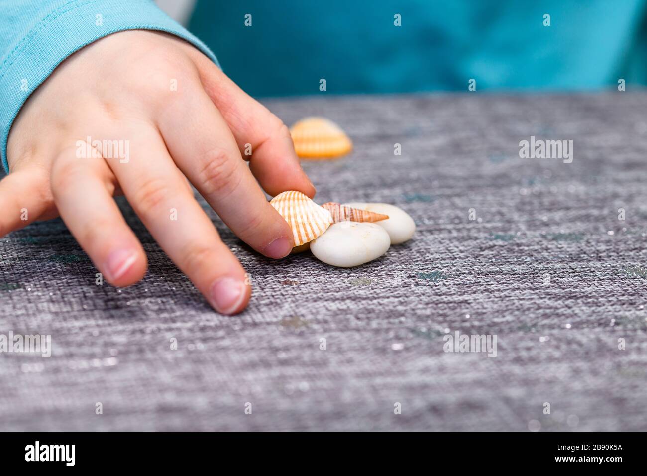 Primo piano di ragazzi di cinque anni che giocano a mano con conchiglie e ciottoli su un tavolo. Ha una camicia turchese e raccogliendo un conchiglia. Foto Stock