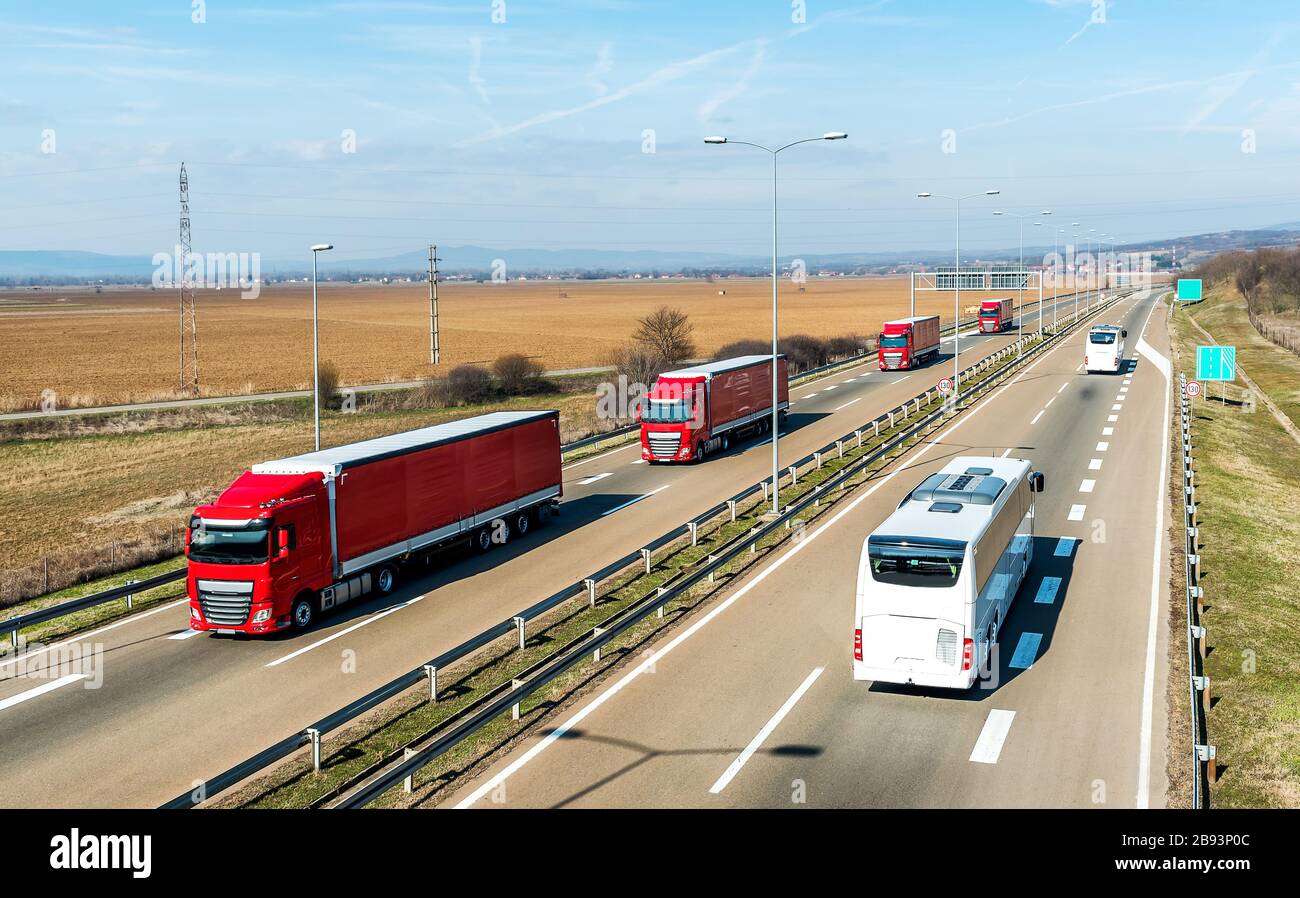 Convoglio di camion di trasporto rossi in linea passando due autobus bianchi su una strada rurale di campagna sotto un bel cielo blu Foto Stock