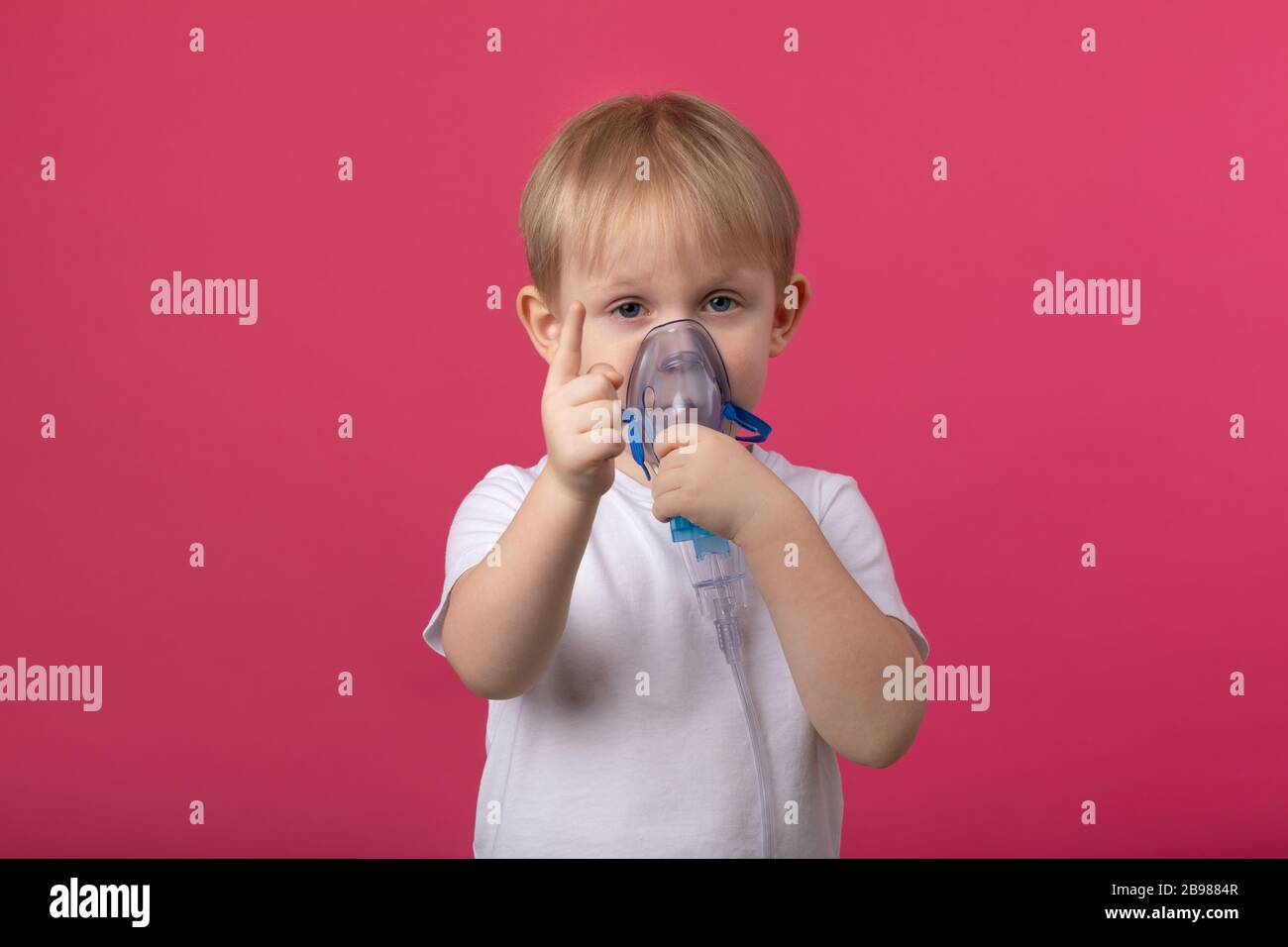 Un bambino biondo con un inalatore in mano attaccato alla bocca punta un dito su uno sfondo rosa chiaro. Studio fotografico per argomenti medici di t Foto Stock