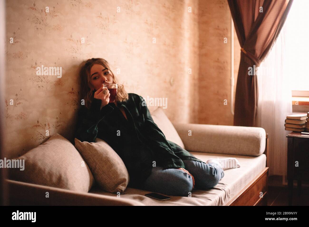 Giovane annoiato donna bere il tè mentre siede sul divano nel soggiorno a casa Foto Stock