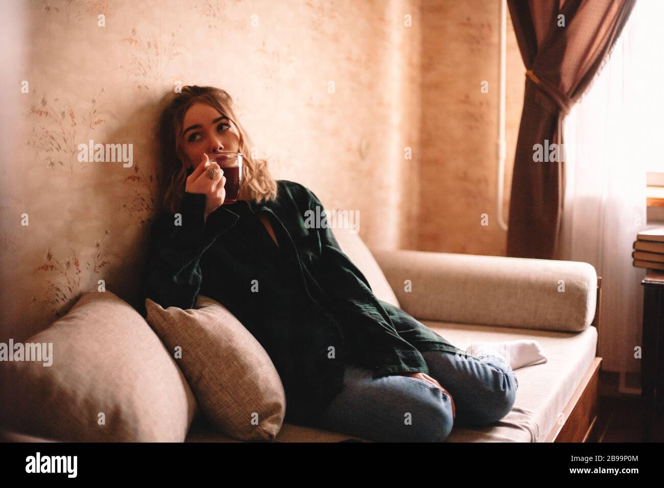 Giovane donna premurosa che beve il tè mentre si siede sul divano nel soggiorno a casa Foto Stock