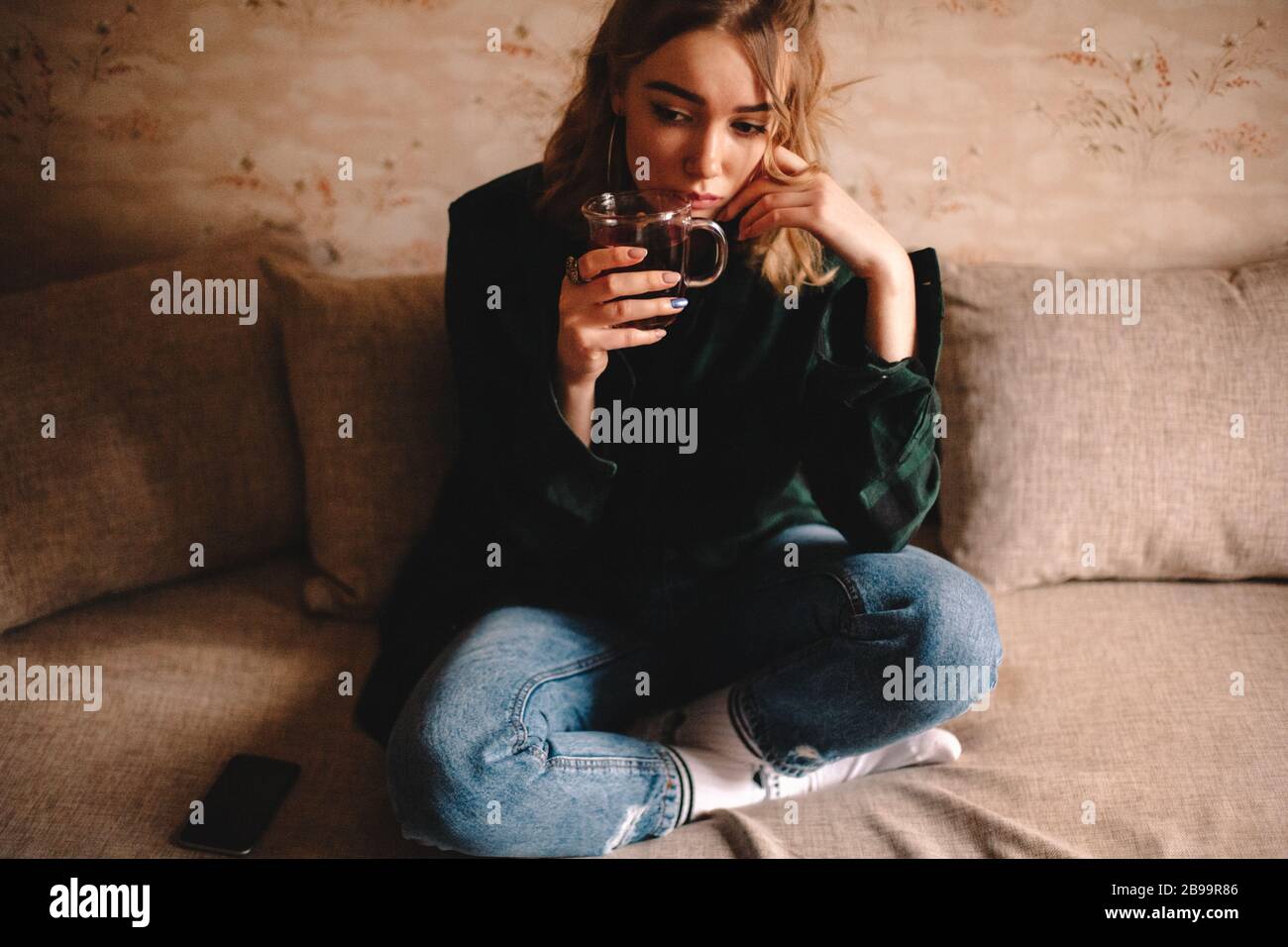 Giovane donna premurosa che beve il tè mentre si siede sul divano a casa Foto Stock
