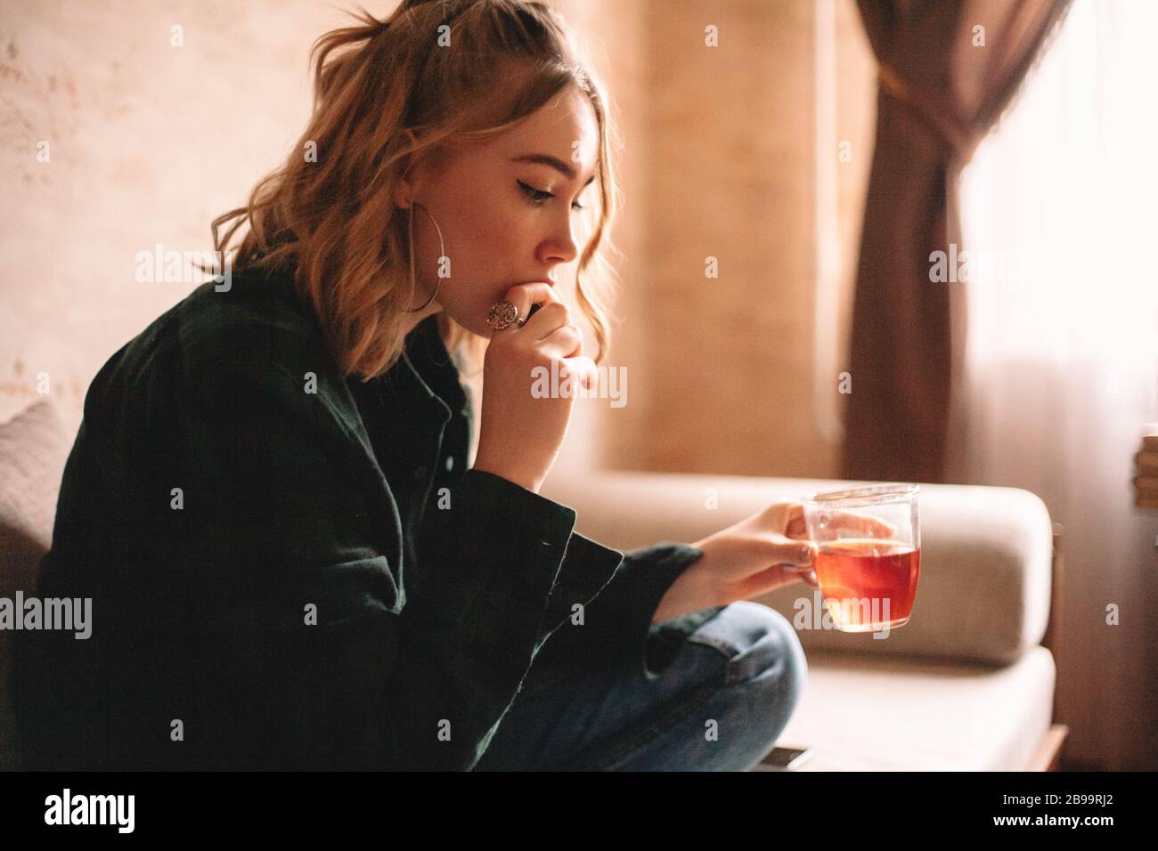 Giovane donna sconvolgente bere tè e mangiare cioccolato mentre si siede sul divano in soggiorno a casa Foto Stock
