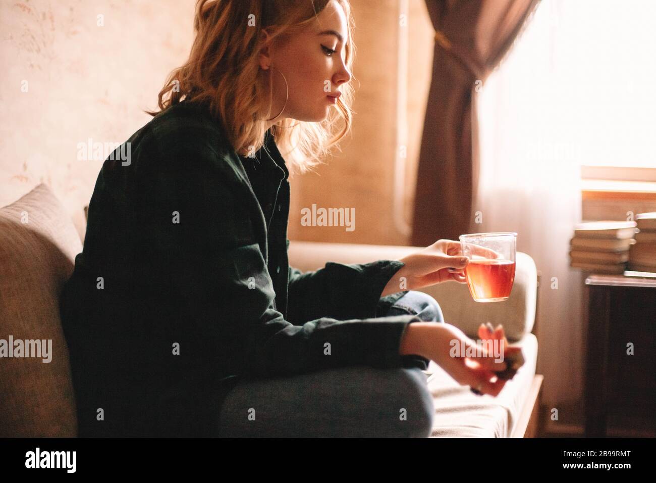 Donna giovane turbata bere tè e mangiare cioccolato mentre si siede sul divano in soggiorno a casa Foto Stock