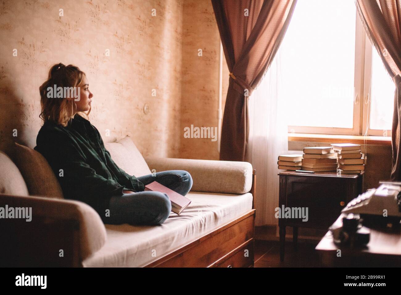 Giovane donna triste che tiene il libro guardando attraverso la finestra mentre siede sul divano in soggiorno a casa Foto Stock