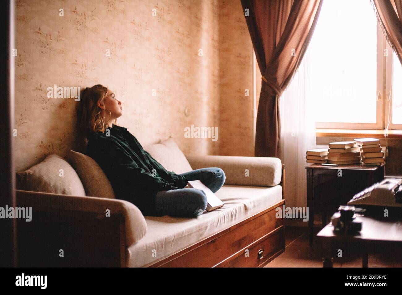 Giovane donna sconvolto tenendo libro guardando attraverso la finestra mentre si siede sul divano in soggiorno a casa Foto Stock