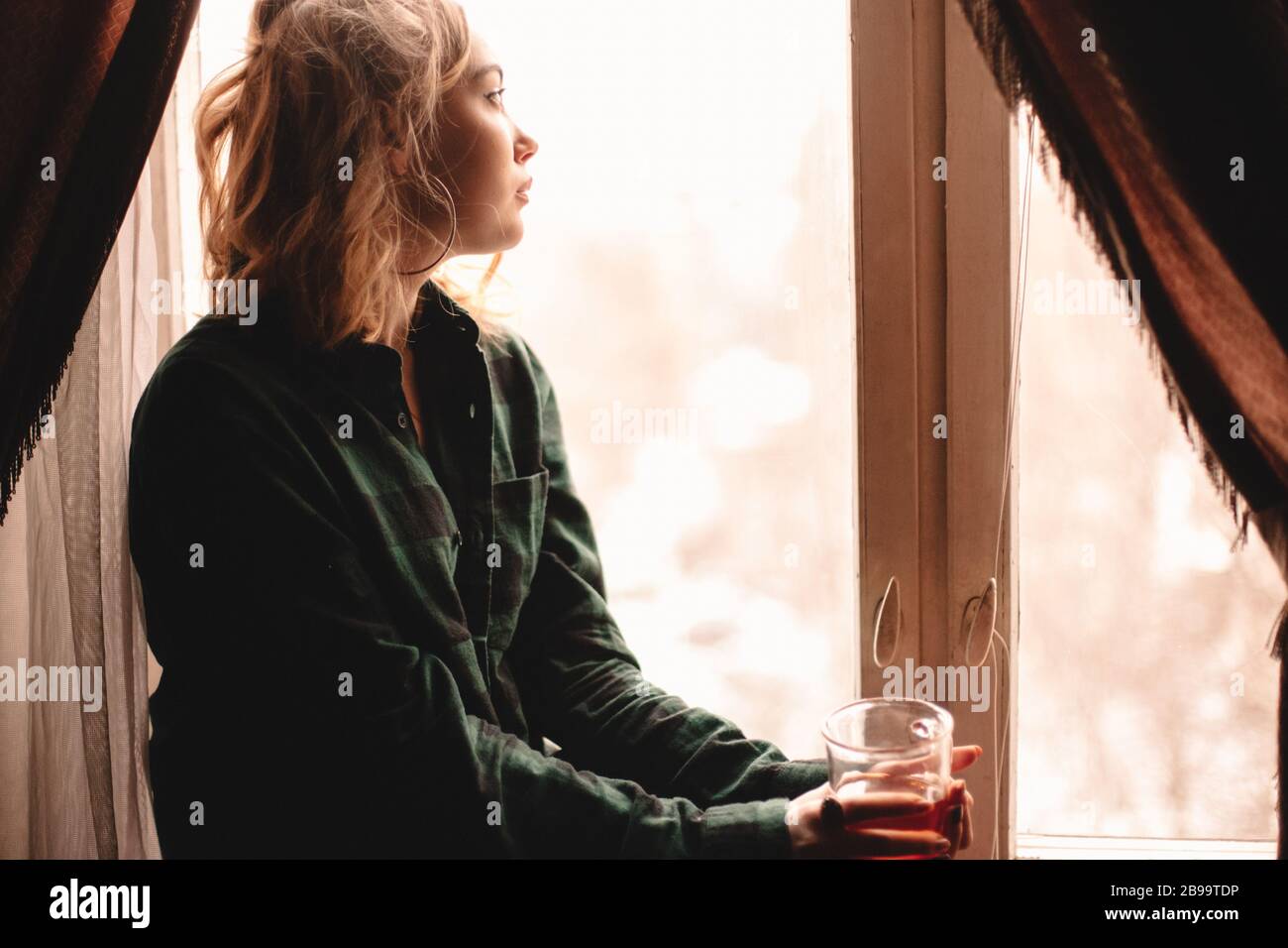 Giovane donna premurosa che beve il tè e guarda attraverso la finestra mentre si siede sul davanzale a casa Foto Stock