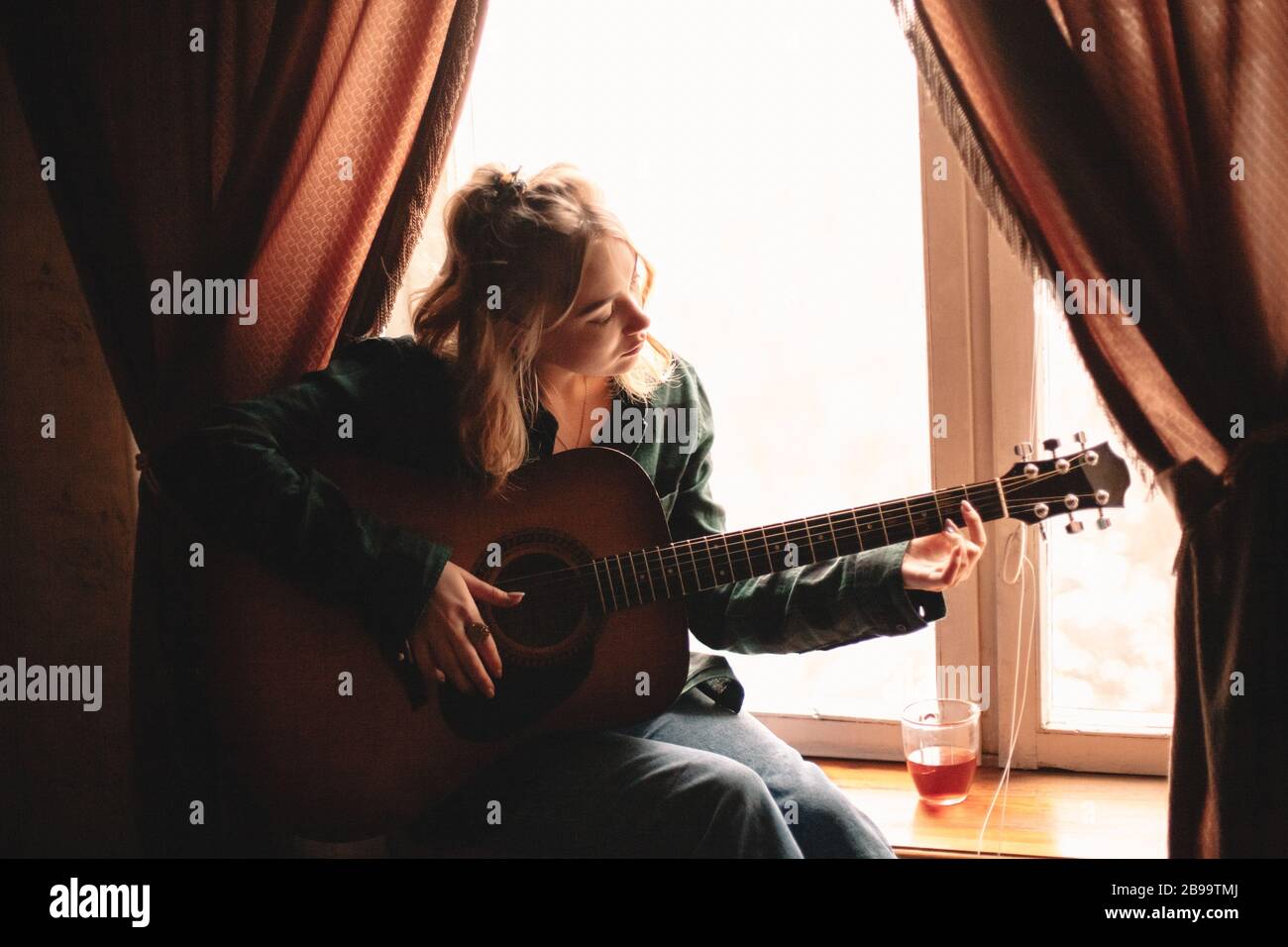 Giovane donna che suona la chitarra mentre si siede su windowsill a casa Foto Stock