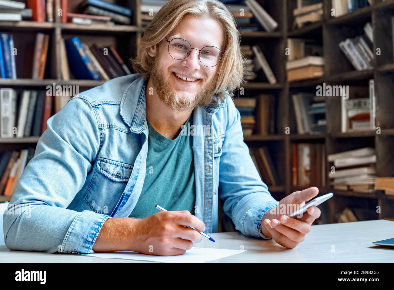 Ritratto di felice giovane allievo sorridente alla scrivania della biblioteca tenere smartphone moderno. Foto Stock