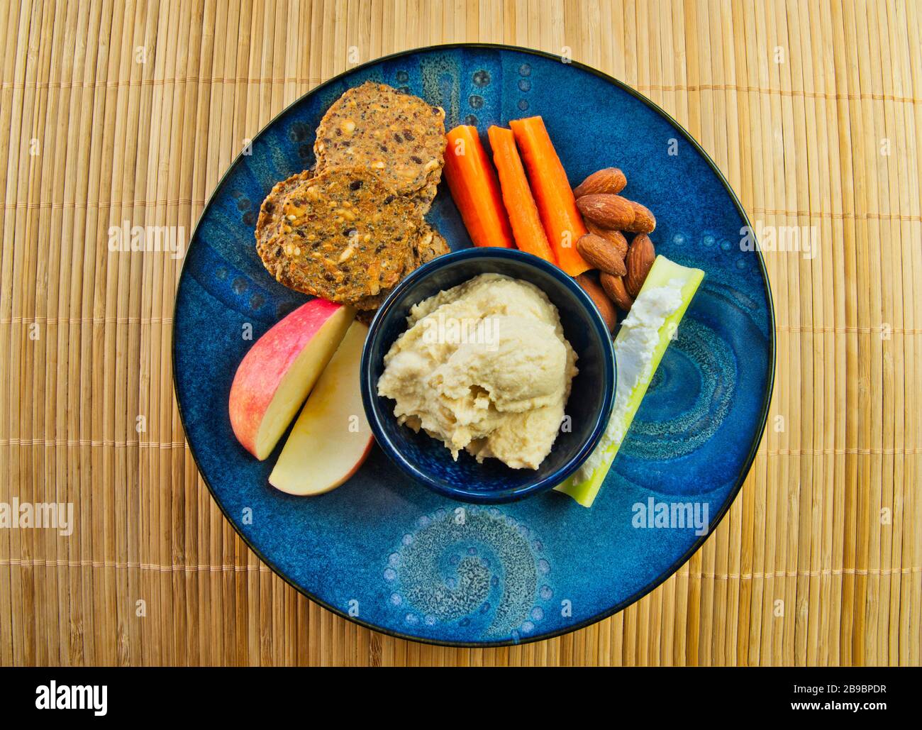 Spuntino o pranzo salutare con hummus, carote, mandorle, sedano, mele e cracker di grano intero su un piatto blu colorato. Foto Stock