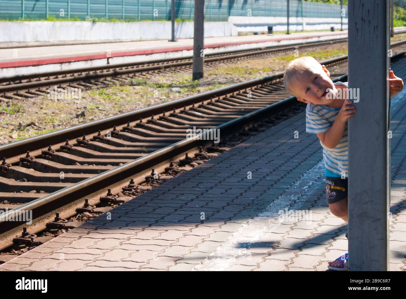 Un piccolo ragazzo dai capelli biondi di di tre anni in una T-shirt e shorts sulla piattaforma sta aspettando il treno e giocando catch-up - sorridendo, ridendo, mostrando la sua lingua. Ricreazione, tropici, vacanza, ferrovia, stazione, estate Foto Stock