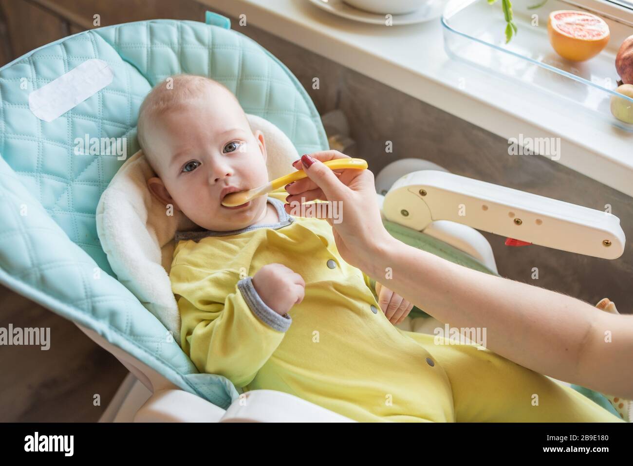 La mamma alimenta la purea di verdure del bambino in un seggiolone. La madre mette un cucchiaio di schiacciamento nella bocca del bambino. Il bambino mangia cibo. Foto Stock