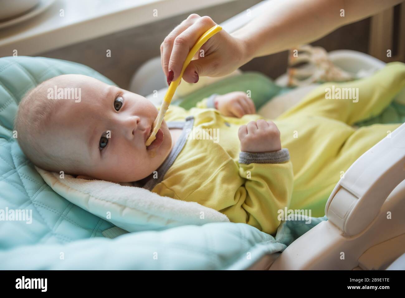La mamma alimenta la purea di verdure del bambino in un seggiolone. La madre mette un cucchiaio di schiacciamento nella bocca del bambino. Il bambino mangia cibo. Foto Stock