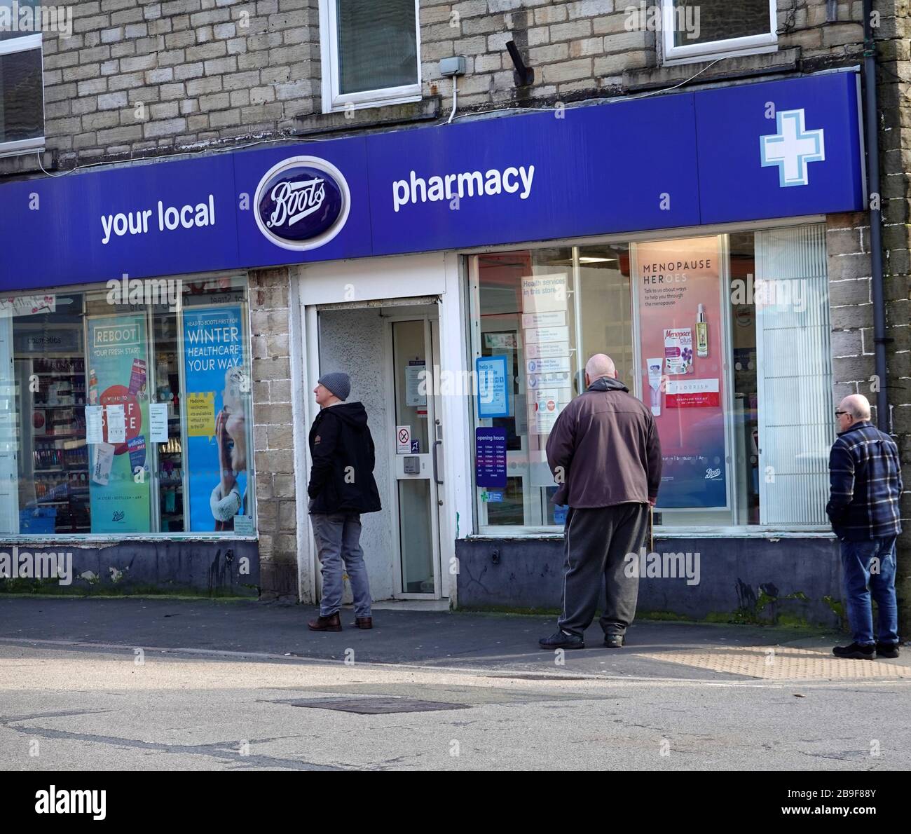 New Mills, Derbyshire. 24 marzo. 2020 tre clienti attendono di entrare in Boots, consentendo solo tre clienti in negozio alla volta. Gli uomini stanno tenendo a parte, anche se non abbastanza due metri come indicato. Le istruzioni del governo sono per la gente rimanere nel paese durante la pandemia del coronavirus coved-19. Le farmacie sono tra quei negozi autorizzati ad essere aperti. Foto Stock