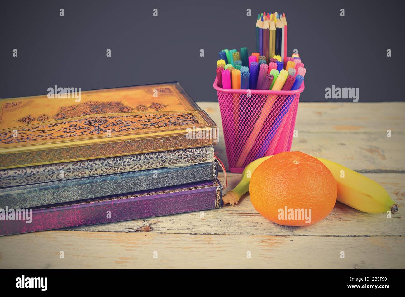 Concetto di pausa pranzo a scuola con banana, tangerine e forniture scolastiche su scrivania in legno, fuoco selettivo. Libri, pennarelli e matite colorate enfatizzando Foto Stock