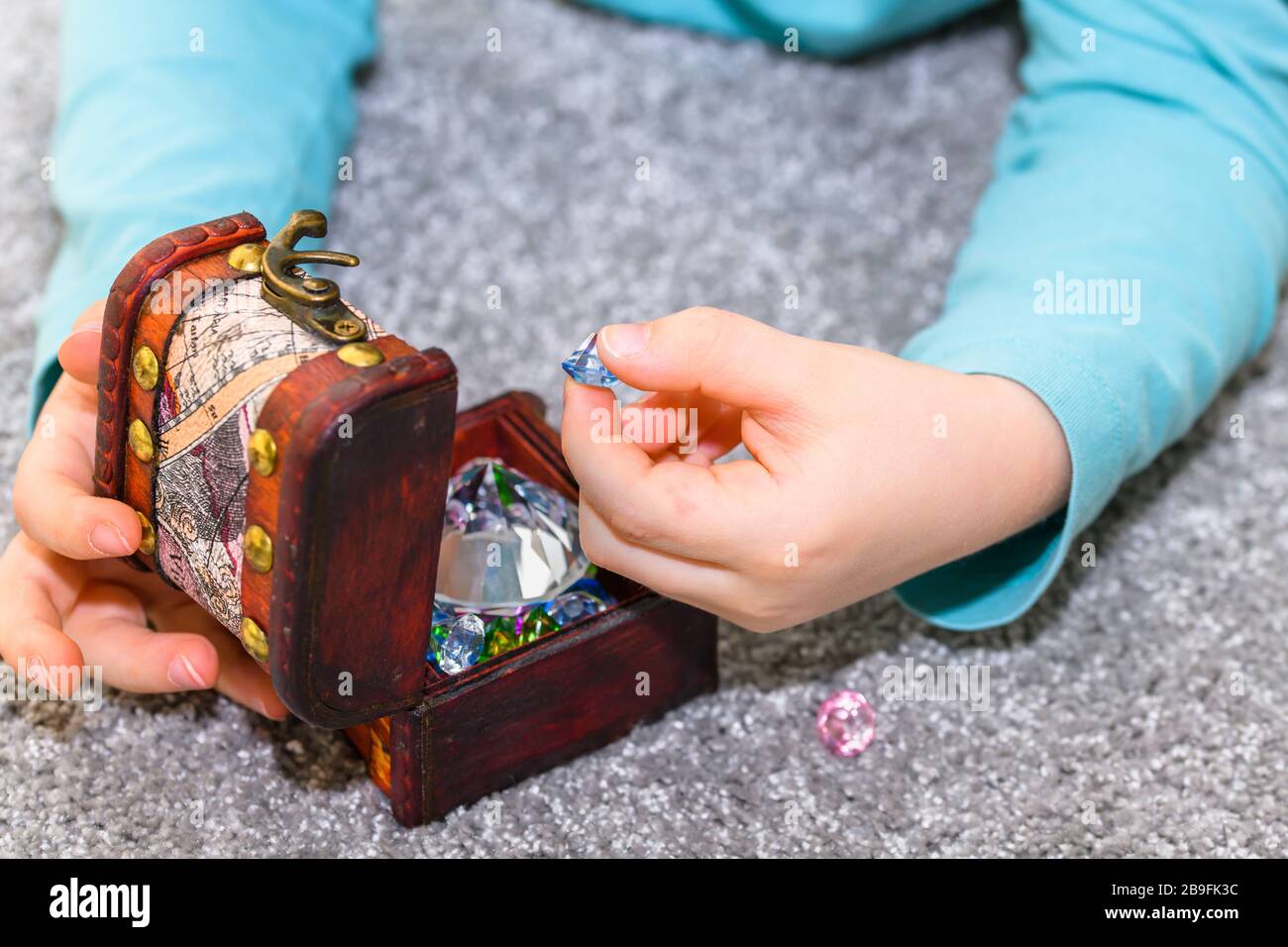 Ragazzo di cinque anni che gioca con una cassa del tesoro piena di gemme multicolori. Il ragazzo ha una camicia turchese e tiene una gemma blu. Foto Stock