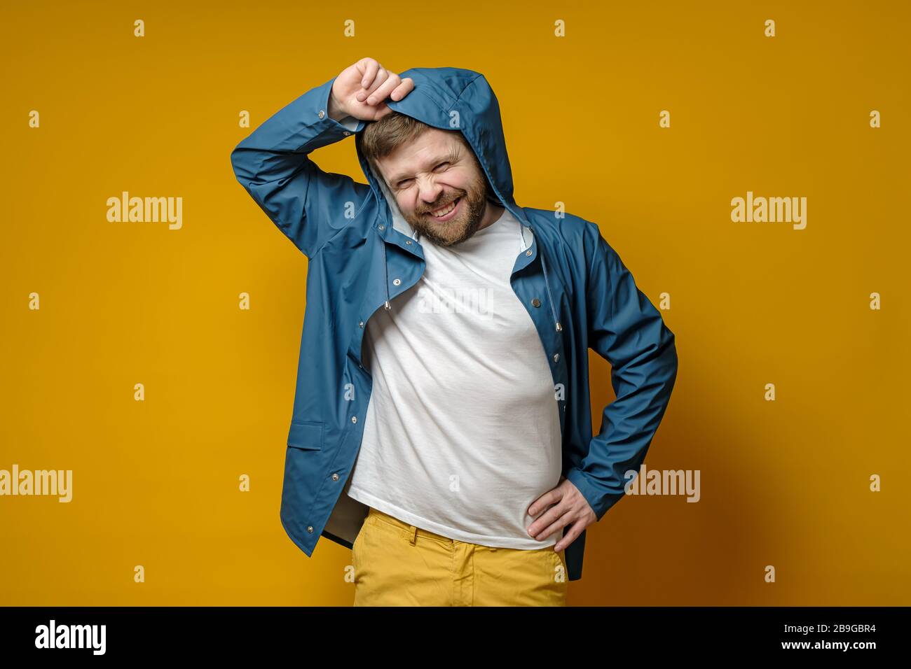 Attraente uomo caucasico bearded in un impermeabile mette su un cappuccio e sorride carino. Isolato su uno sfondo giallo. Foto Stock