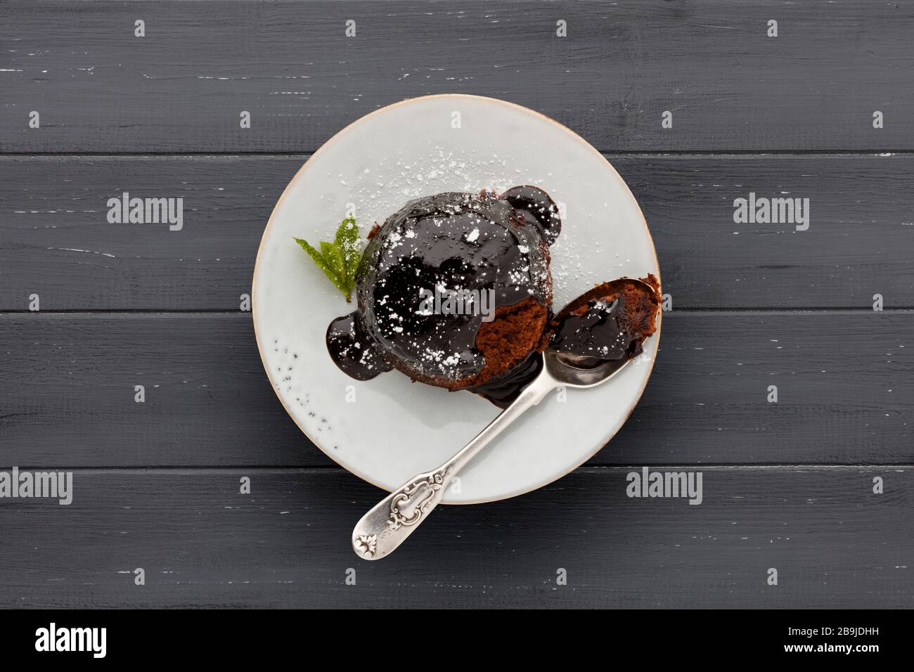 Un singolo pan di Spagna al cioccolato, con una salsa di cioccolato appiccicosa, su un piatto bianco con un cucchiaio, sparato su una superficie di legno grigio scuro con spazio per c Foto Stock